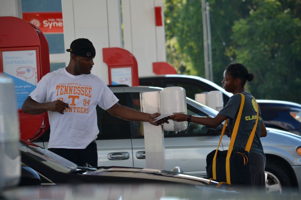 A person handing someone a book near a gas station