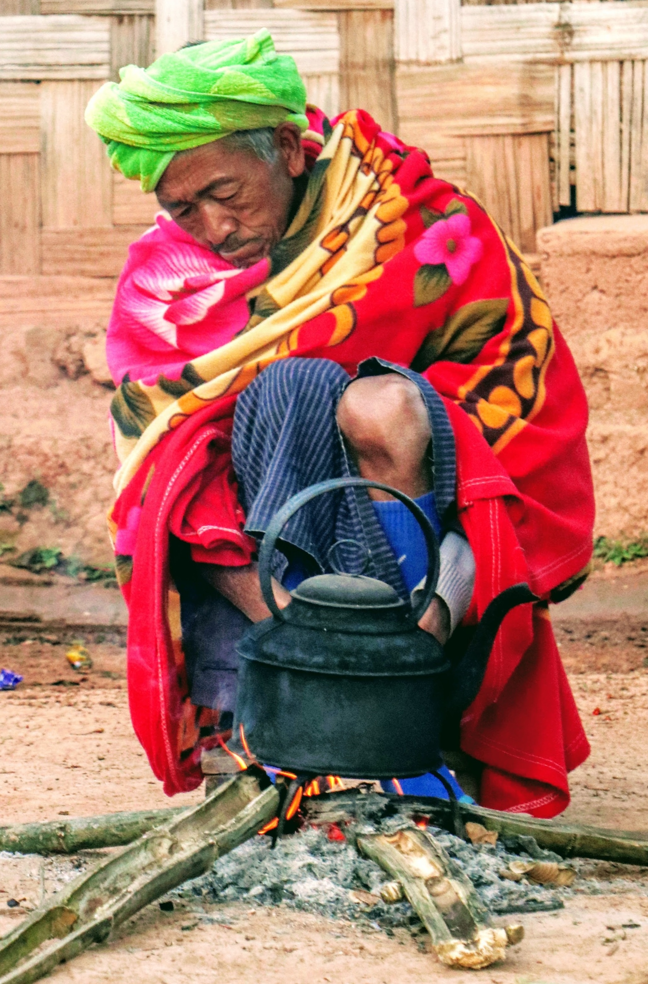 Old Burmese villager in bright green headwrap and red robe aslweep by his outdoor fire while waiting for tea pot to boil.