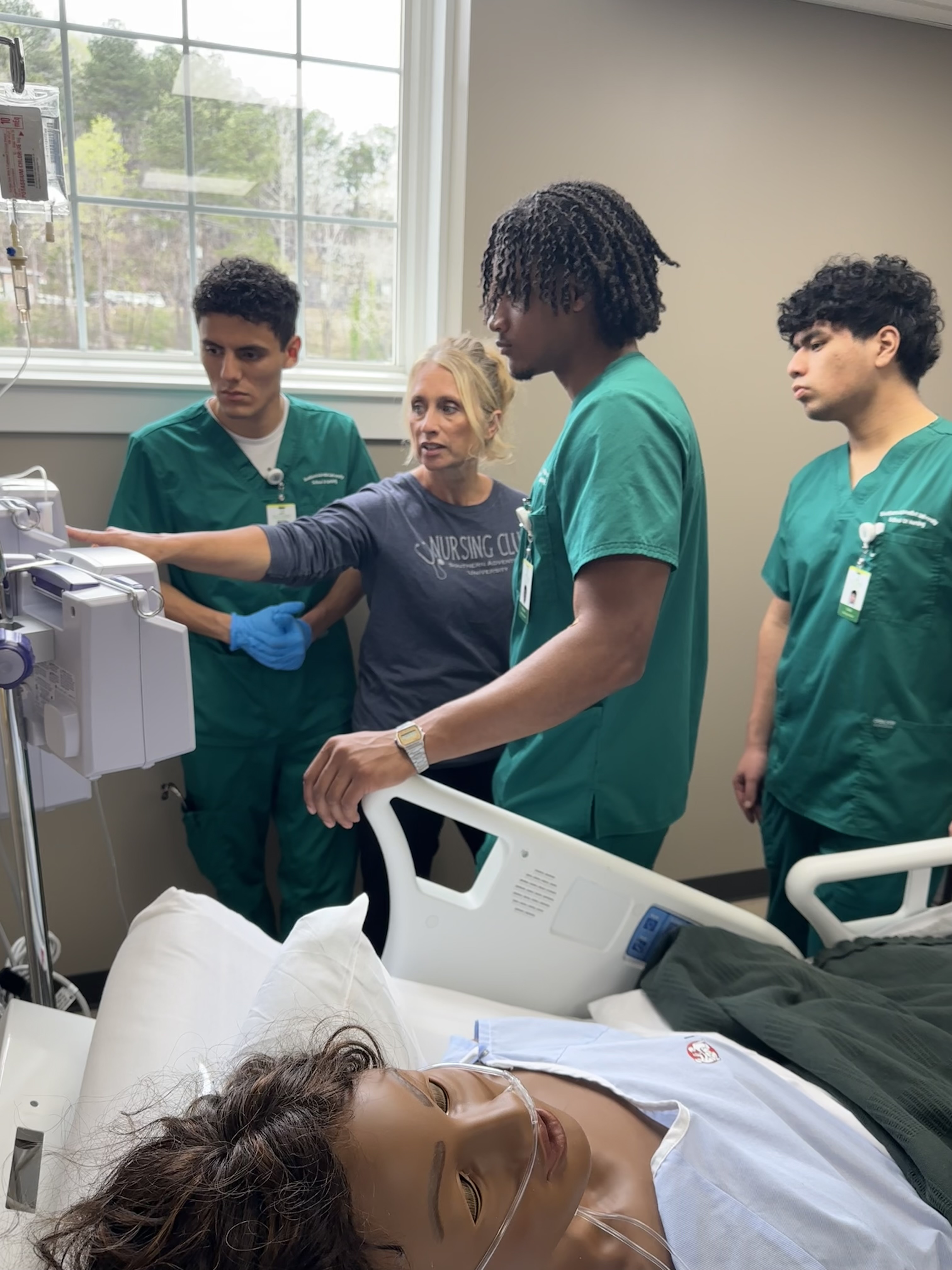 Several students observe as a professor indicates the IV pump next to a manikin patient.