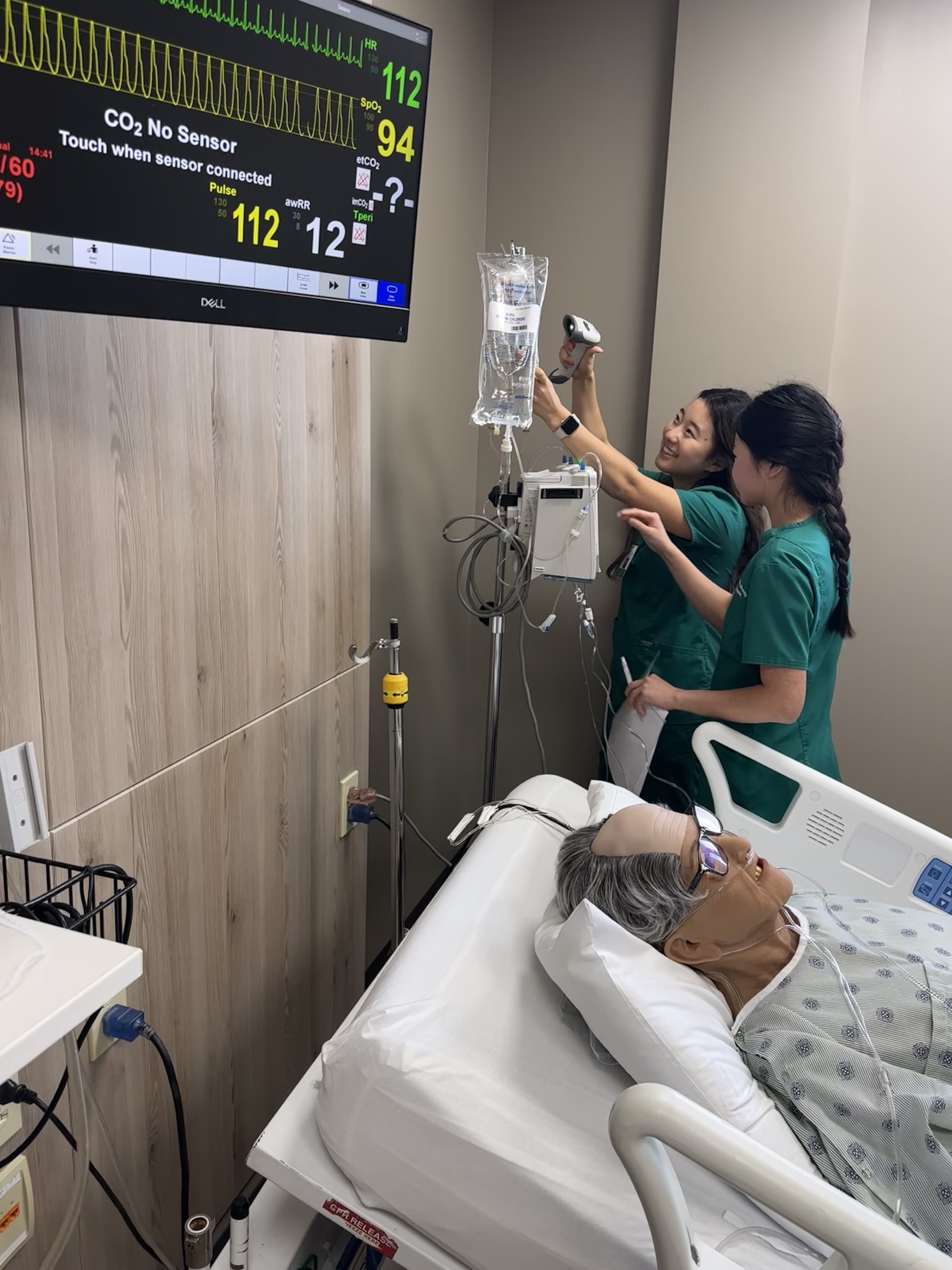 Two students practice operating an IV pump for an elderly manikin patient.