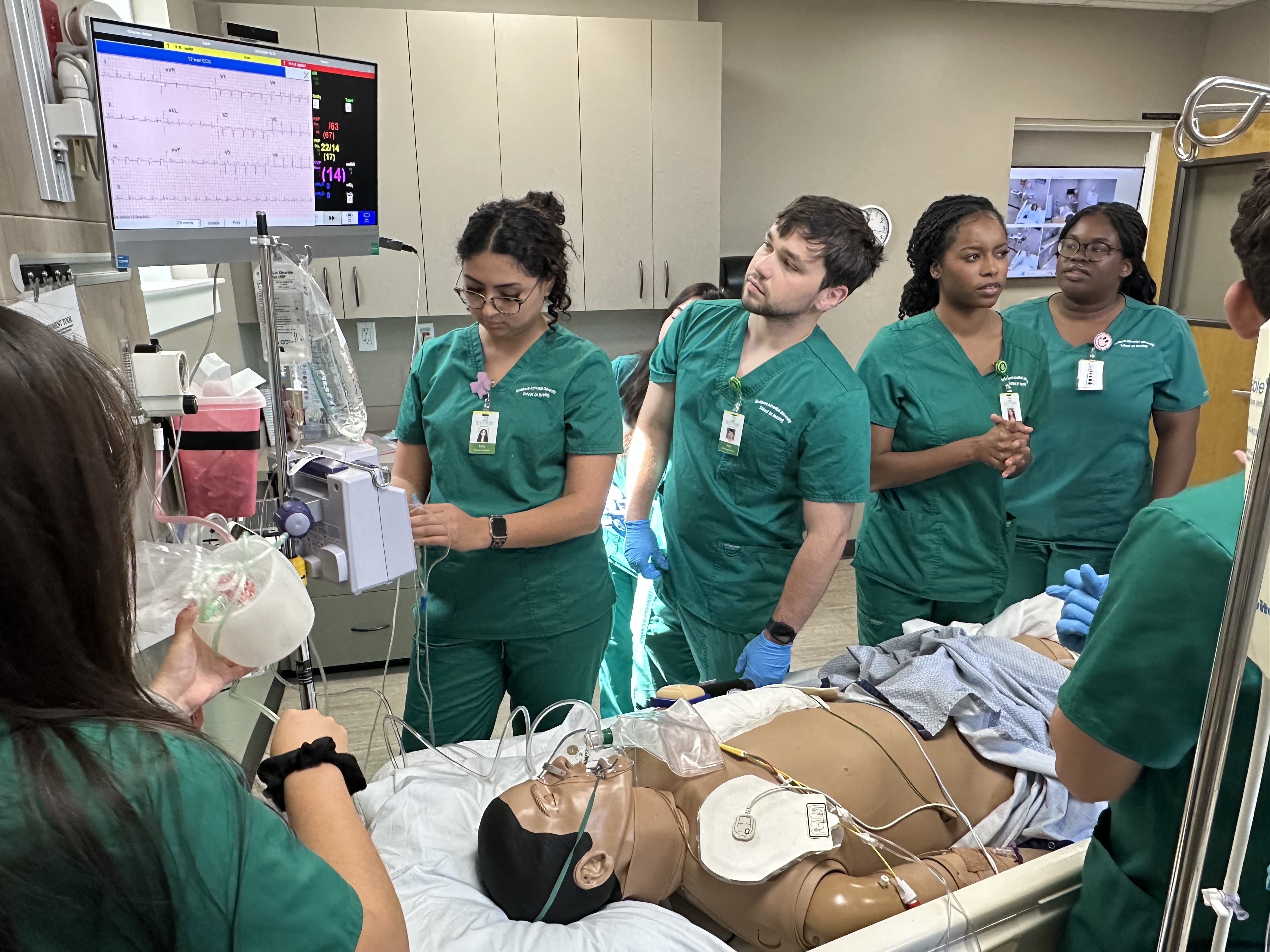 Six students surround a manikin patient's bed. The patient is wearing an oxygen mask.