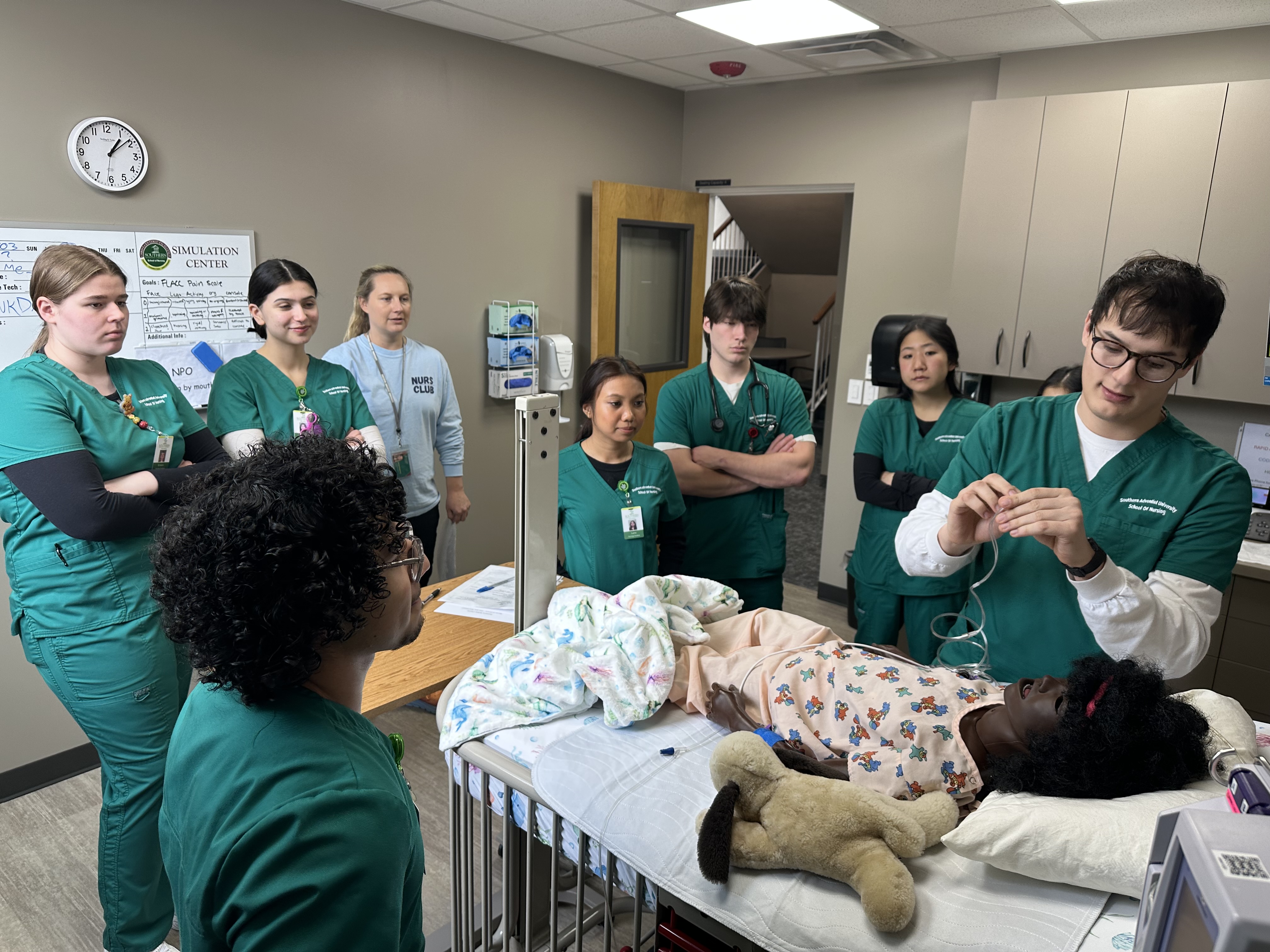 Seven students and a professor watch as a student cares for a child manikin patient.