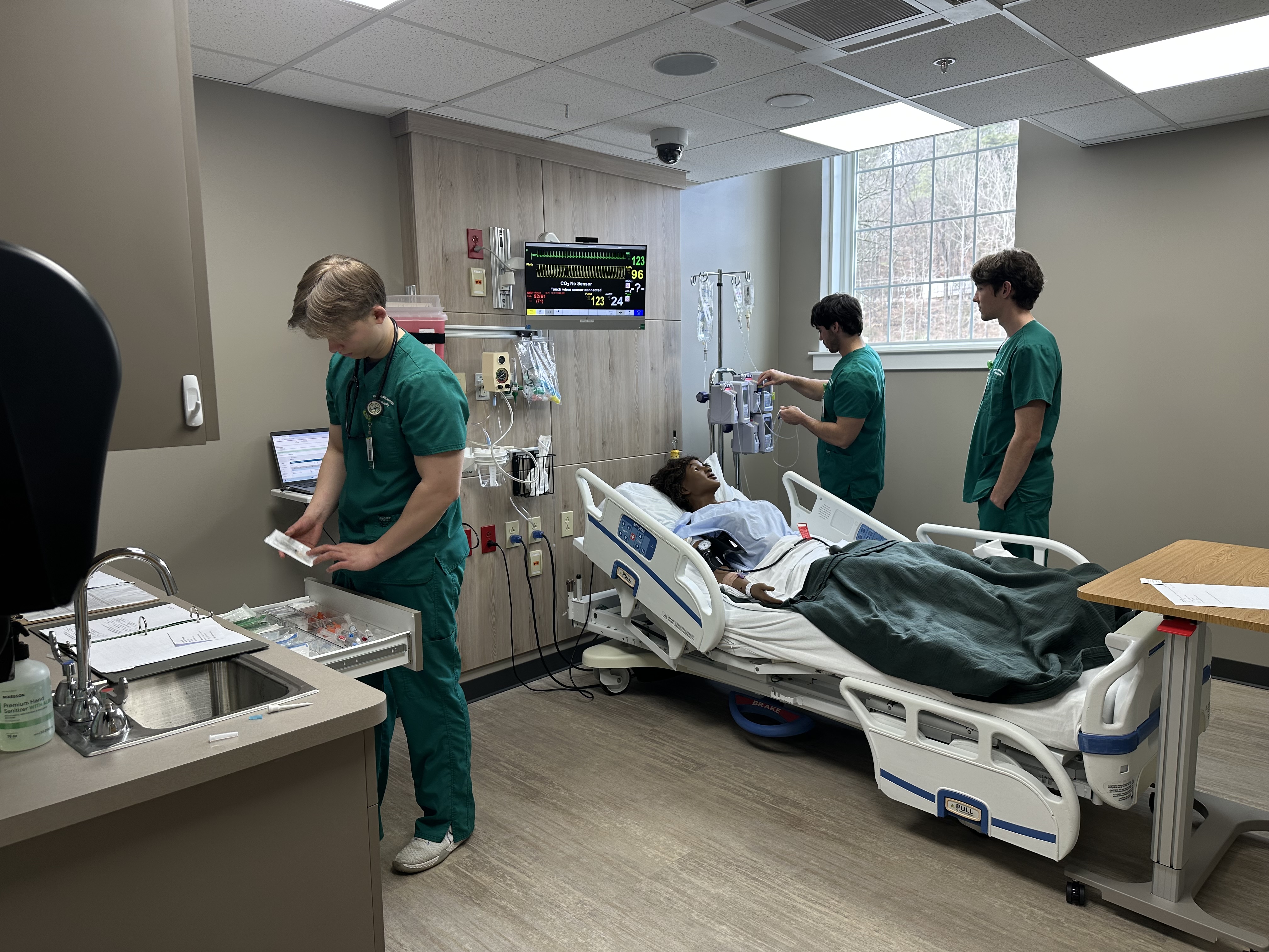 Three students do various tasks in simulation hospital room.