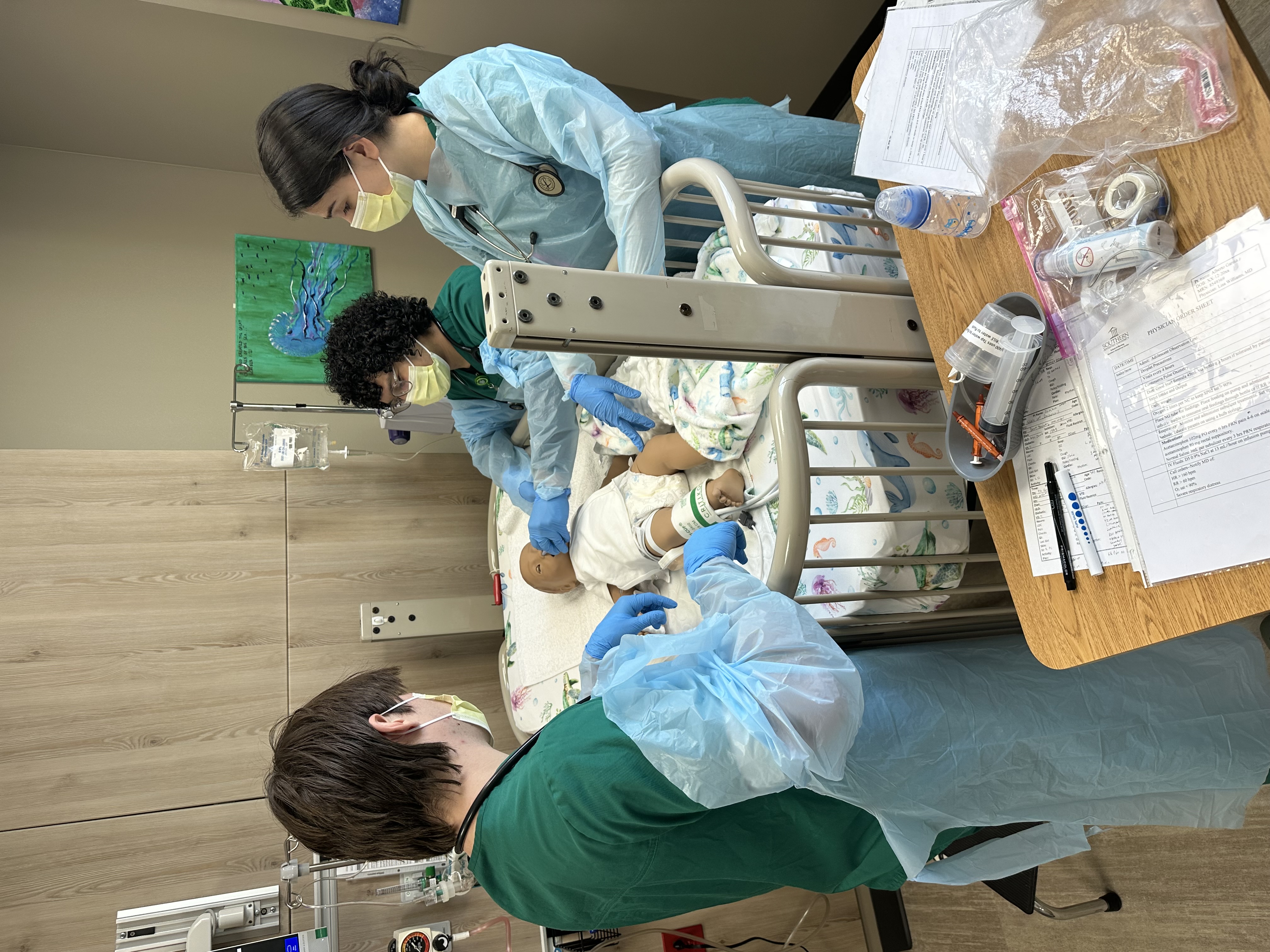 Three students in masks and surgical gowns care for a baby manikin.