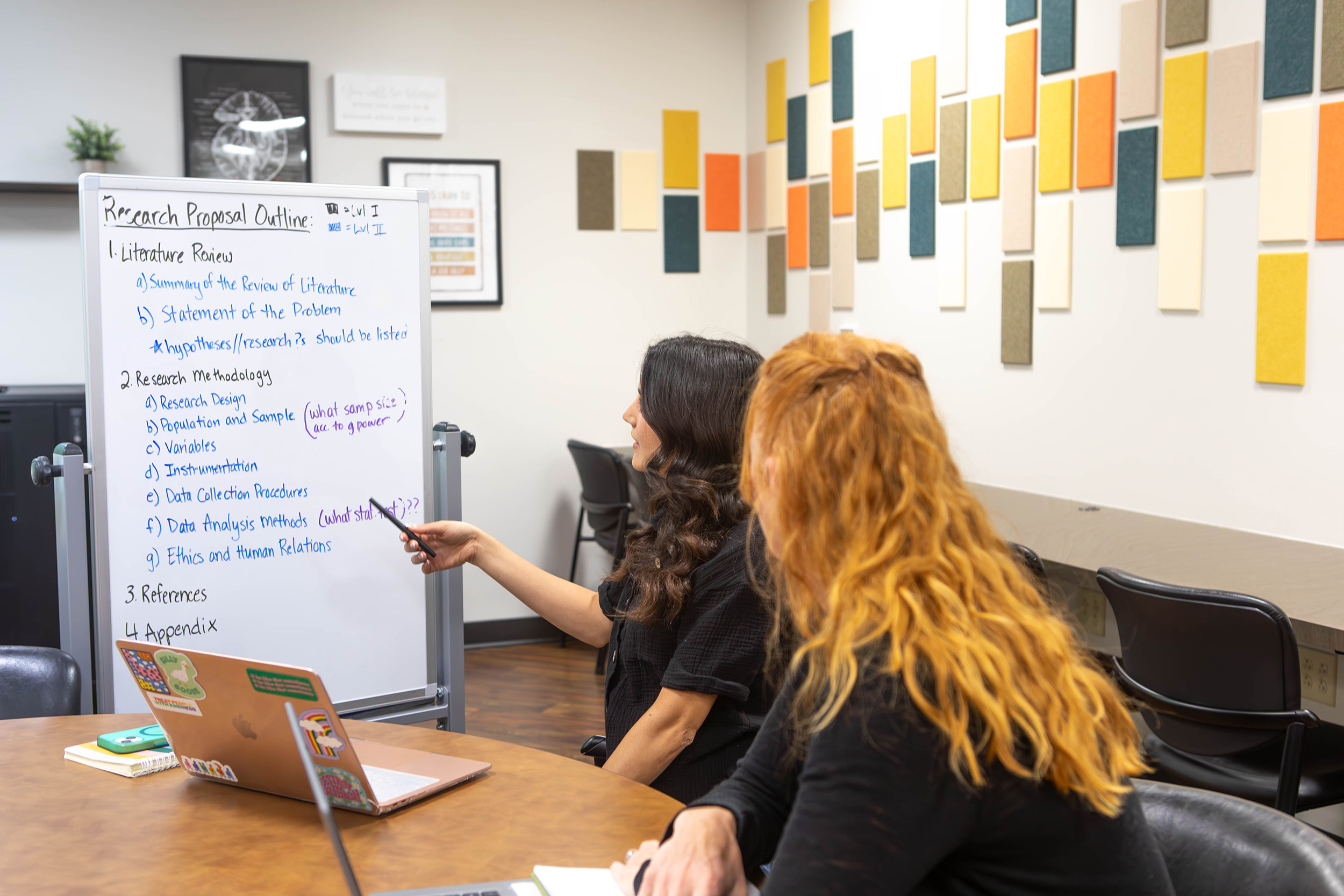 Two students sitting in front of a whiteboard discussing a research proposal outline.