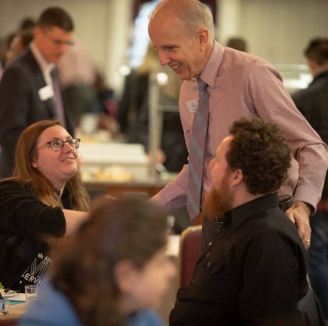 Group of men and women gathered at a table, greeting one another.